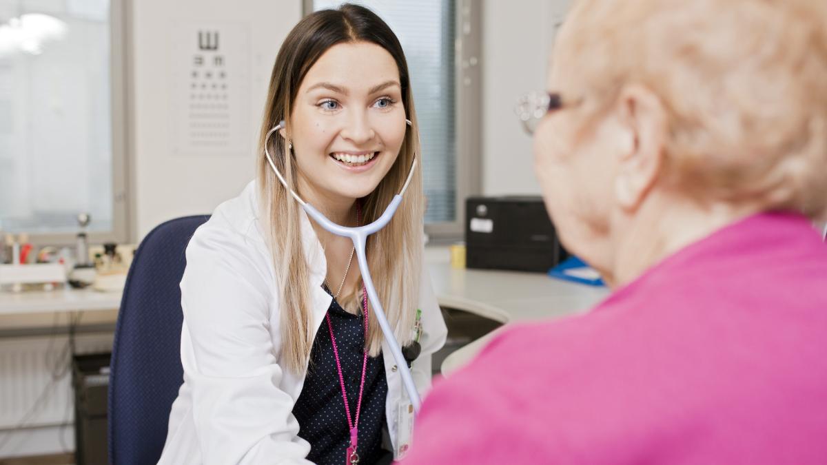 Lääkäri hymyilee ja juttelee vanhemman henkilön kanssa tutkimushuoneessa.