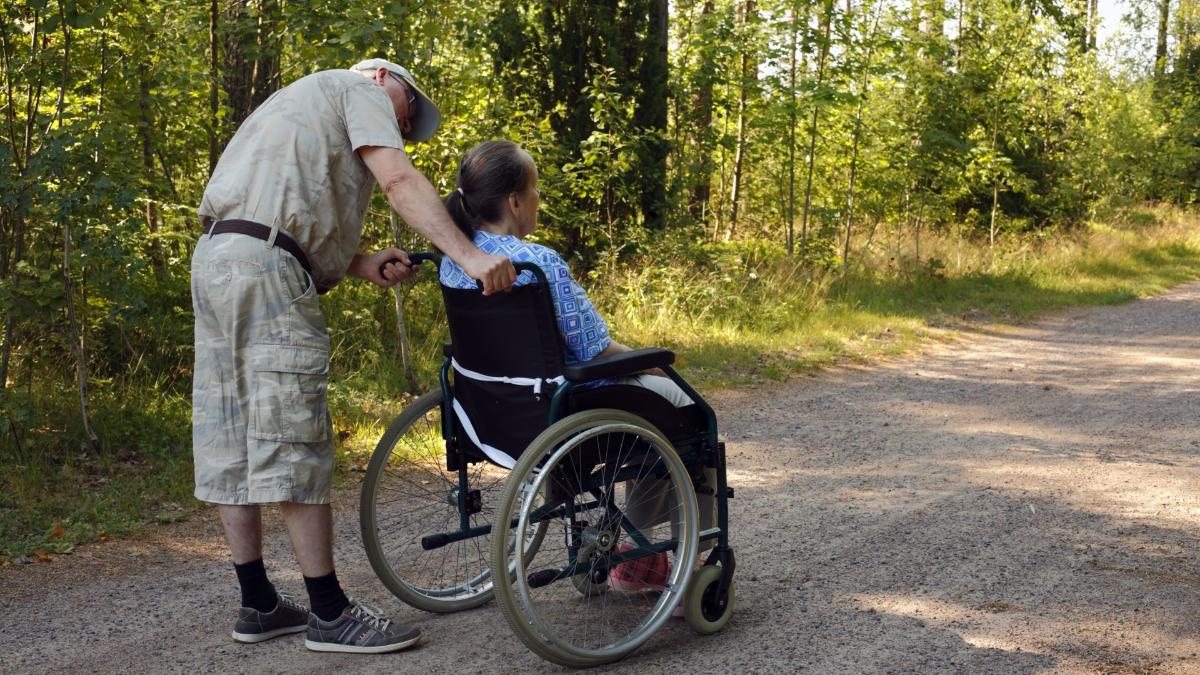 Ikäihminen pyörätuolissa ja toinen ikäihminen pyörätuolin takana luonnon keskellä.