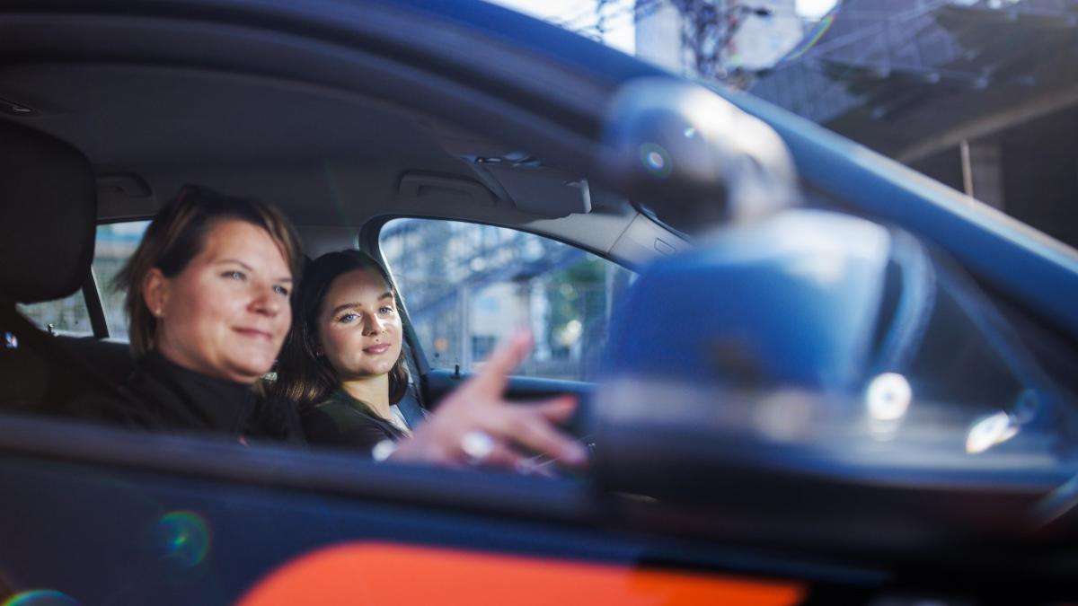 Ajo-opettaja osoittaa peruutuspeiliä autokoulun oppilaalle autossa.