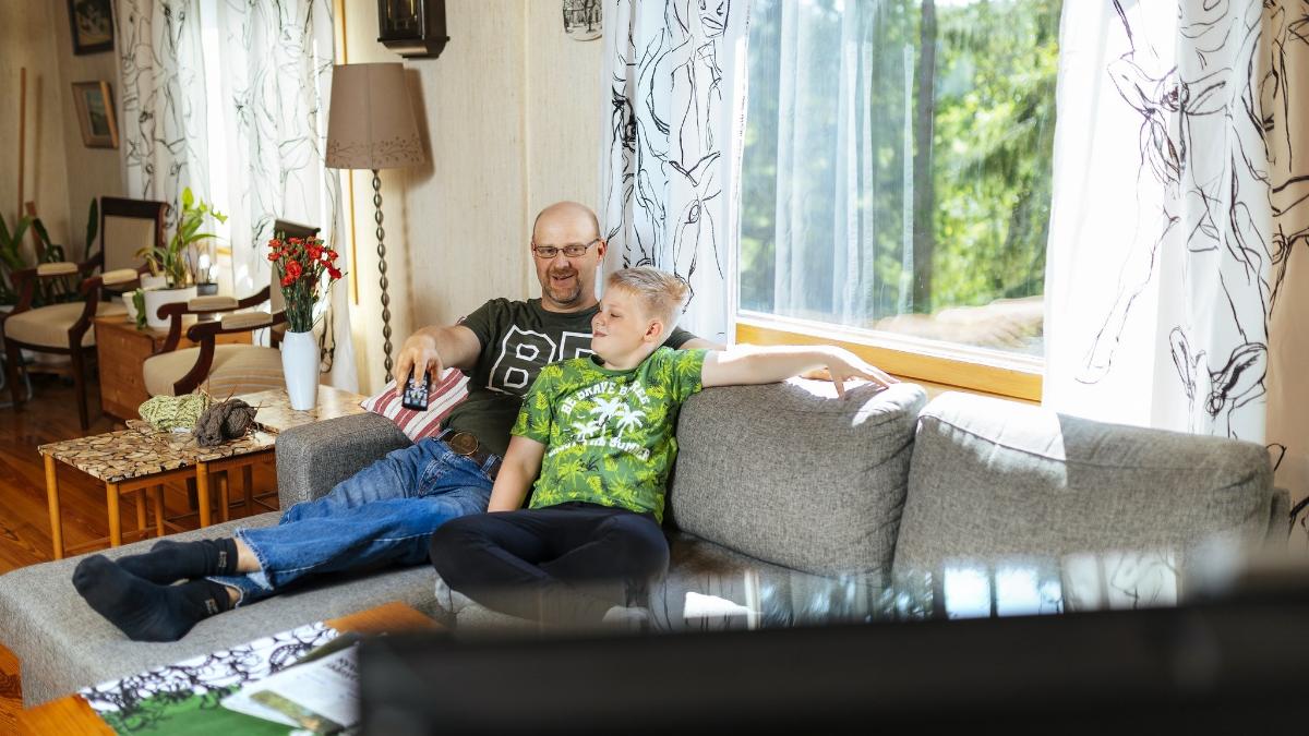 Isä ja lapsi istuvat kotisohvalla ja katsovat televisiota kotonaan.