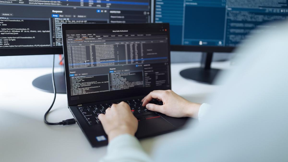 Kannettava tietokone ja kaksi siihen kytkettyä näyttöä, ihminen kirjoittaa koodia kannettavalla tietokoneella.