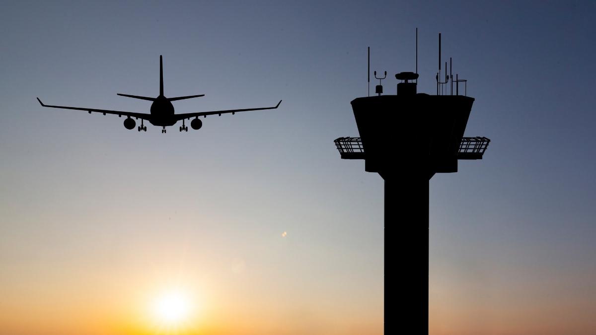 Lentokone lentää lennonjohtotornin lähellä, molemmat näkyvät siluetteina auringonlaskua vasten.