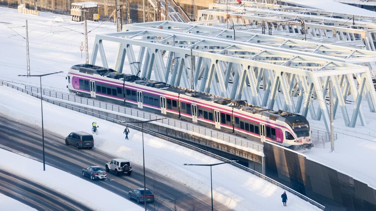 Lähijuna, kävelijöitä ja autoja Pasilan asemalla lumisessa maisemassa.