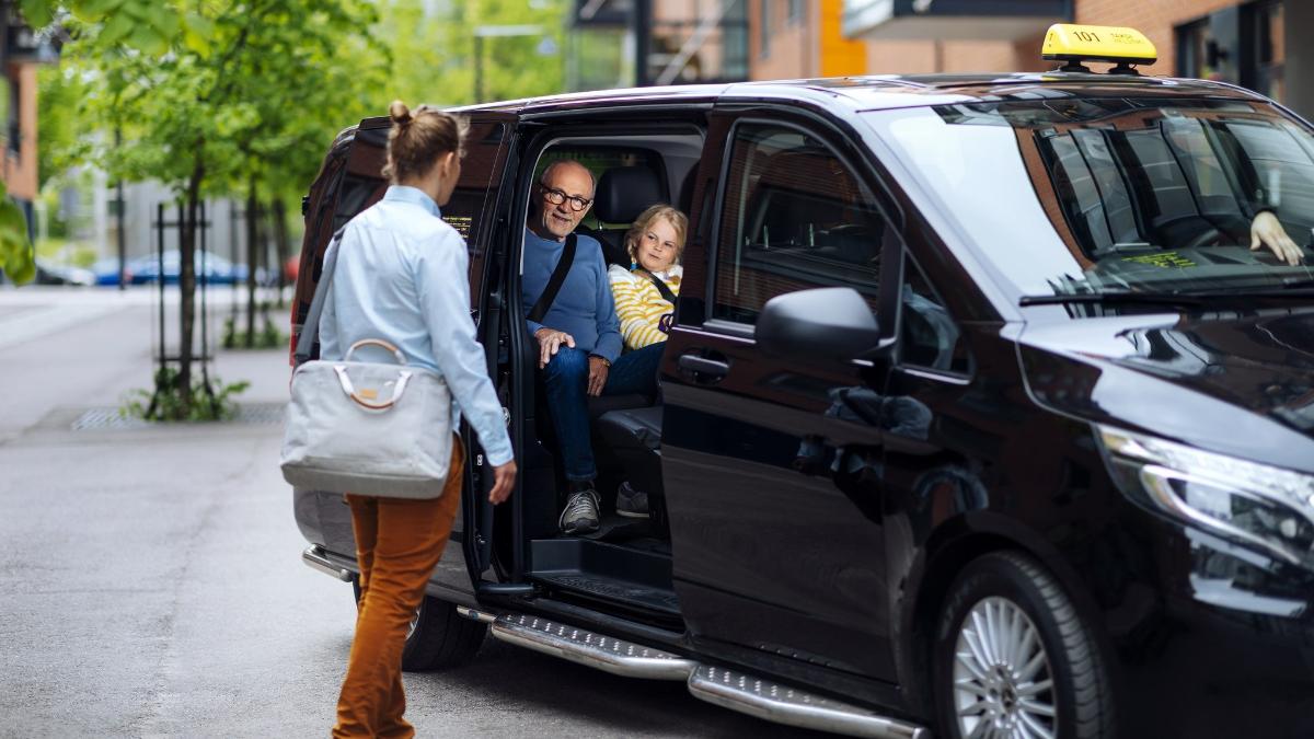 Kaksi matkustajaa tilataksissa ja yksi matkustaja nousemassa kyytiin kesäisessä kaupunkimaisemassa.