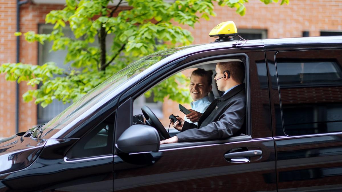 Asiakas istuu etupenkillä taksinkuljettajan vieressä ja maksaa taksimatkan puhelimella.