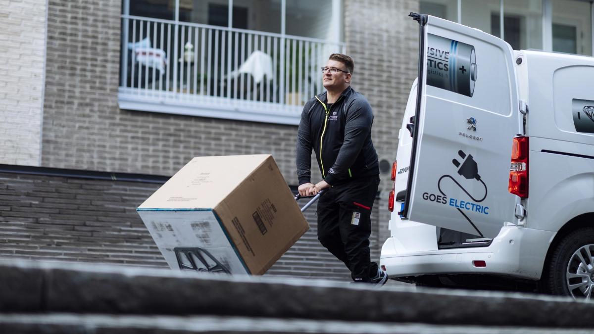 Mies kuljettaa pakettia kärryillä pakettiautosta asuintalon pihalla.