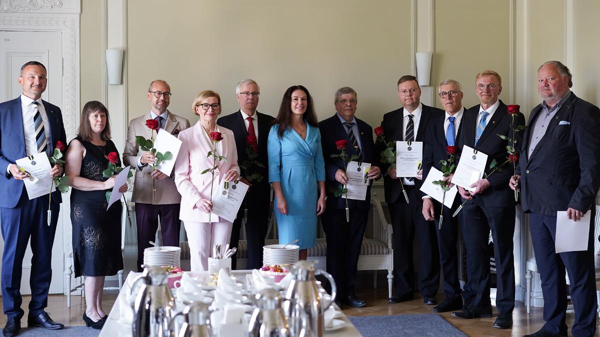 Tio medaljörer poserar tillsammans med ministern. Medaljörerna håller i röda rosor, ett diplom och en medalj. I bildens förgrund syns de tårtbitar som offrats.
