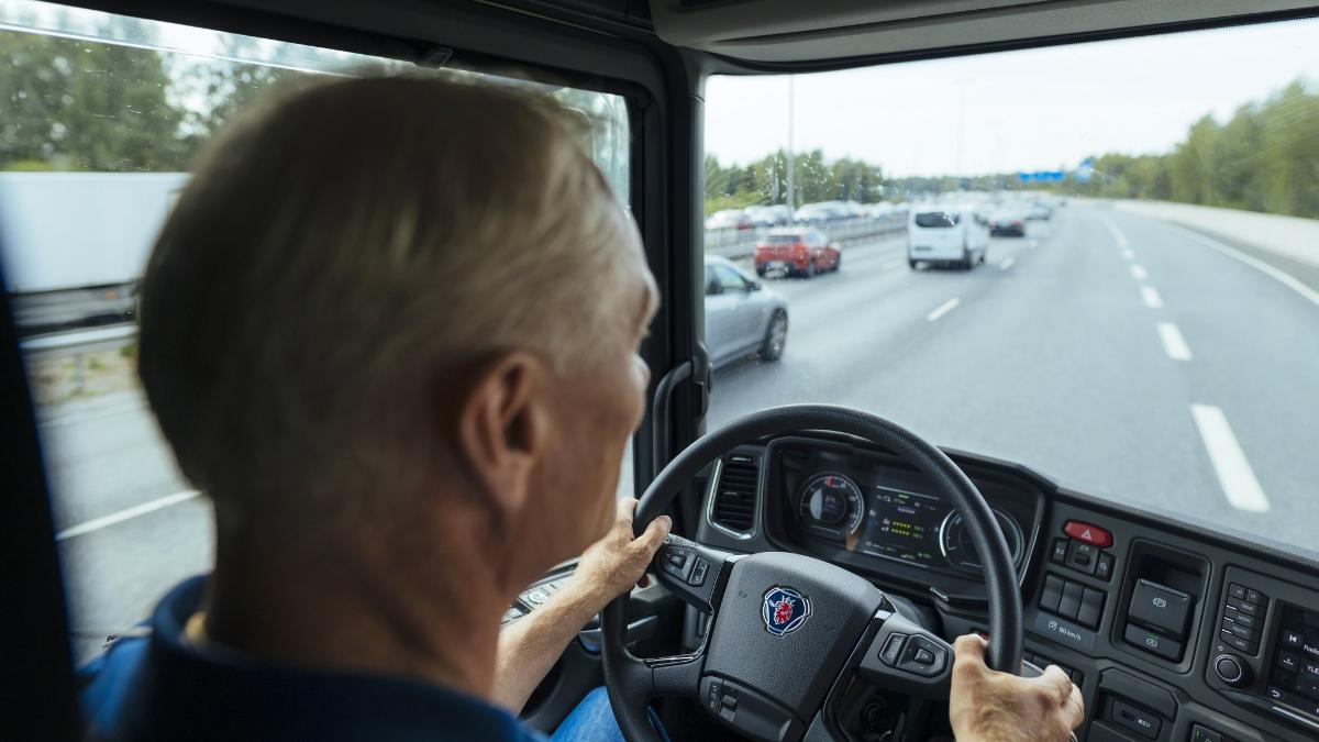 Harmaahiuksinen mies rekan ohjaamossa ajamassa rekkaa.