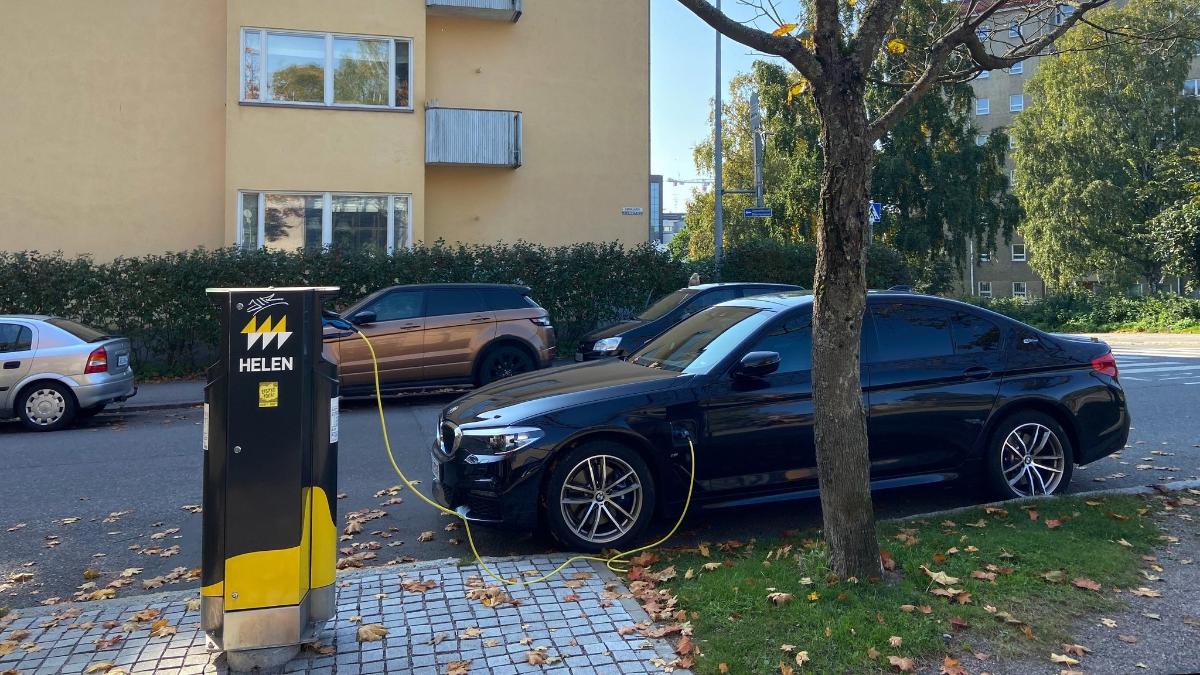 Musta sähköauto latauspisteellä kadun varressa. Taustalla muita autoja ja keltainen talo.