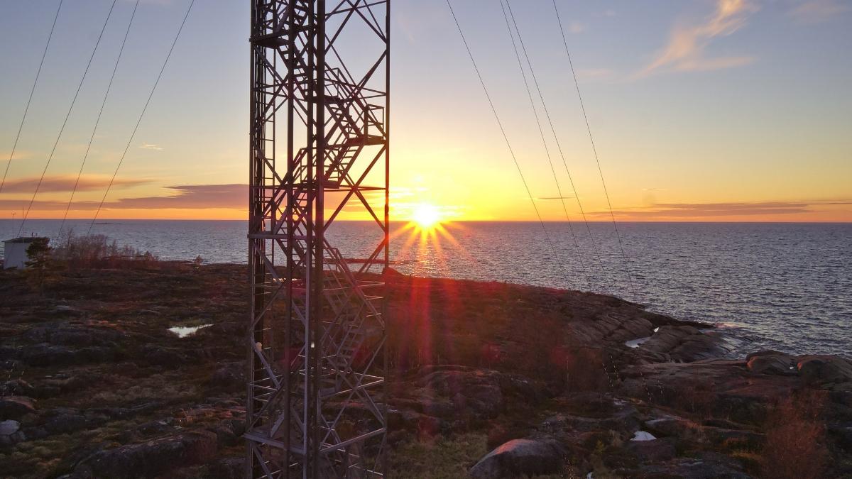 Kuvassa näkyy maston meren rannalla ja taustalla auringonlasku.