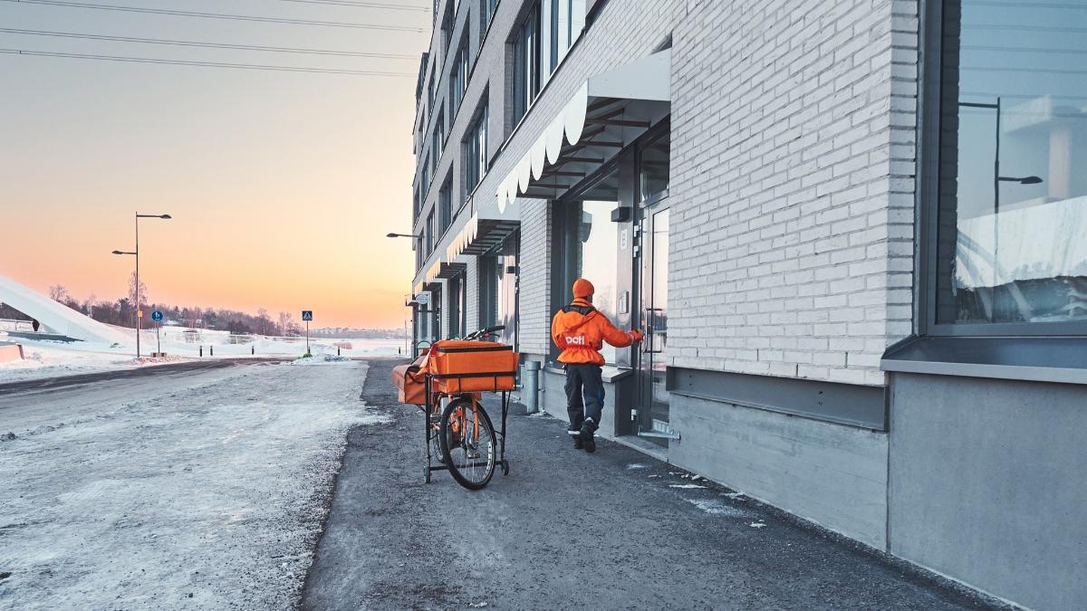 Posteljooni menossa sisään kerrostalon rappukäytävään lumisessa kaupunkimaisemassa.