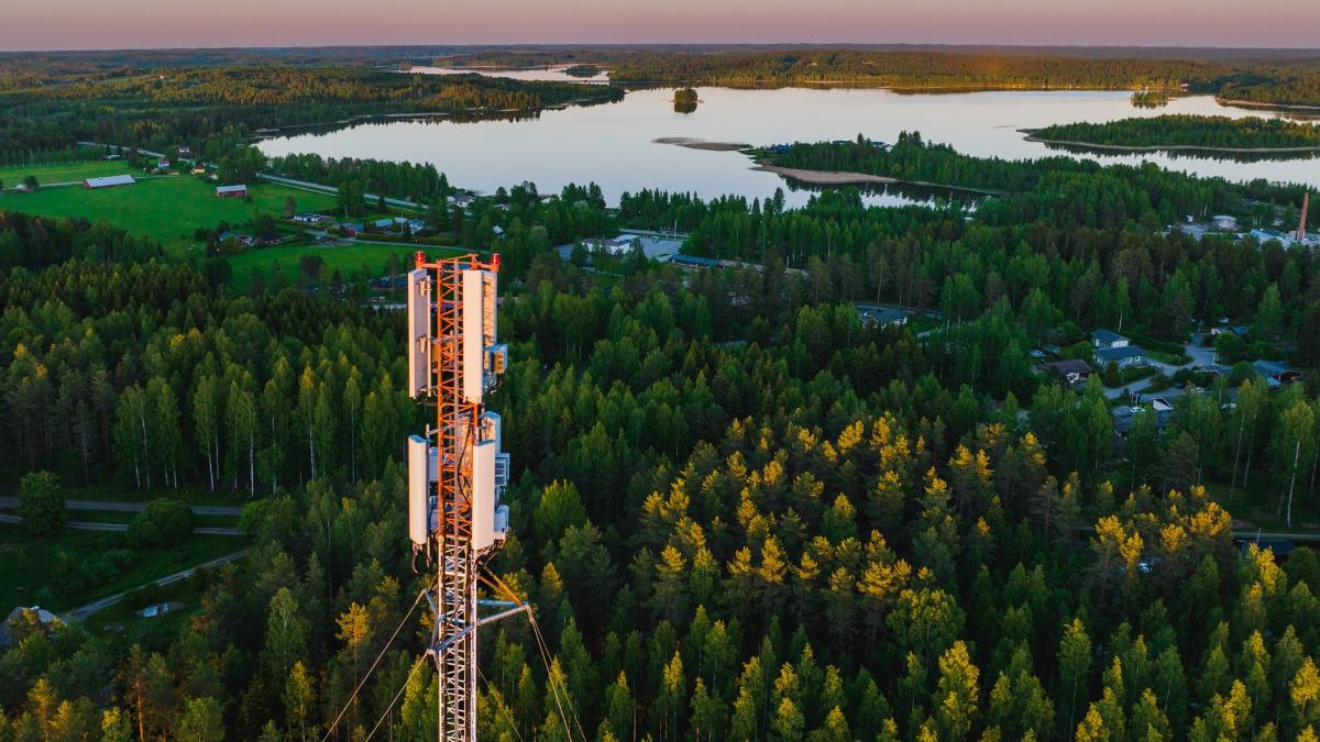 Etualalla tietoliikennemasto. Taustalla vihreää metsää, peltoja ja vesistöä.