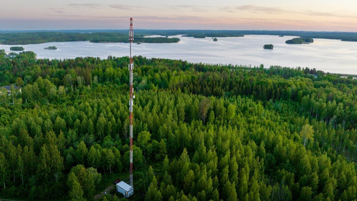 Etualalla tietoliikennemasto vihreän metsän keskellä. Taustalla järvimaisema ja taivasta.