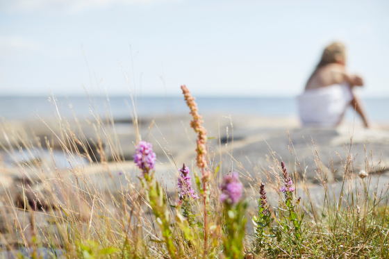 Pyyhkeeseen kääriytynyt henkilö istuu rantakivellä, etualalla kukkia ja heinää.