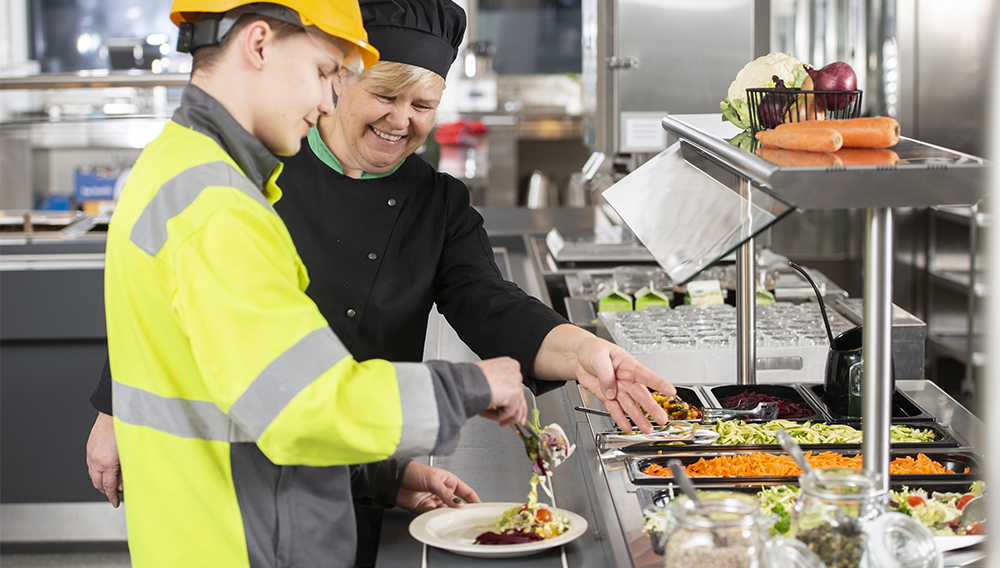 ammattikoulun ruokalassa poika ottaa linjastolta ruokaa ja keittäjä neuvoo vieressä