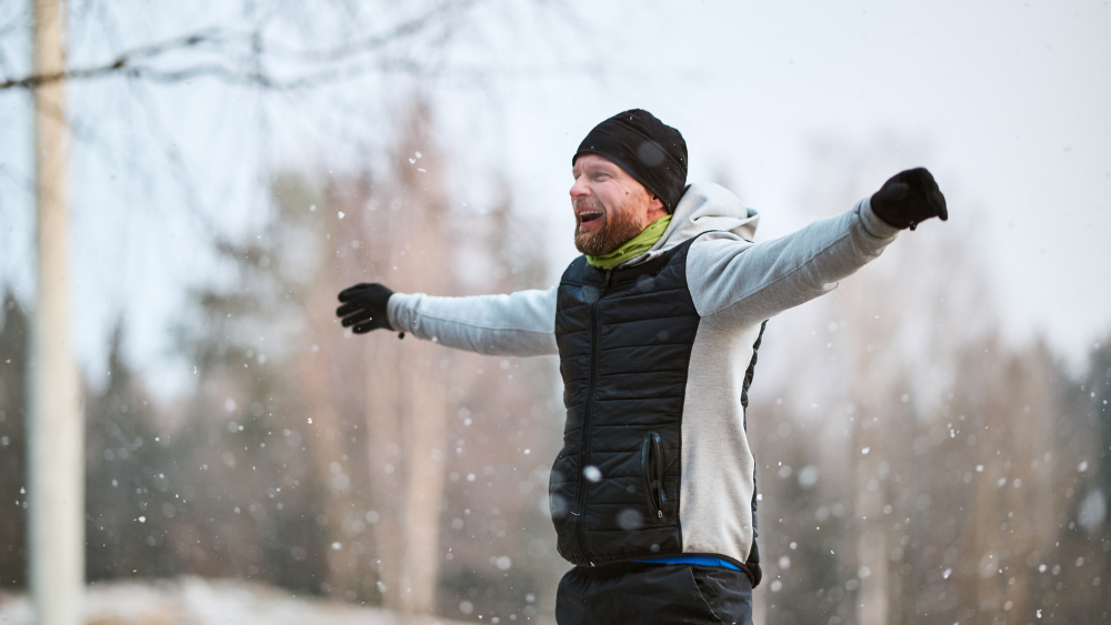 Mies seisoo urheiluvaatteissa kädet vaakatasossa pienessä lumisateessa.