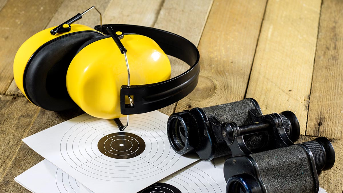 Keltaiset kuulosuojaimet, mustat kiikarit ja maalitaulu puutason päällä.