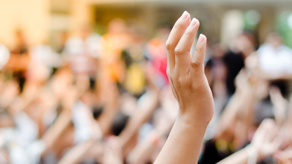 Kuvituskuva. Pystyssä oleva käsi pyytää puheenvuoroa. Taustalla epätarkkana iso joukko ihmisiä.