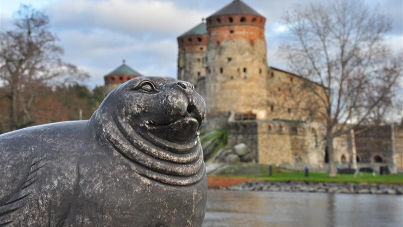 Kuvituskuva: etualalla saimaannorppa-patsas, taustalla Olavinlinna.
