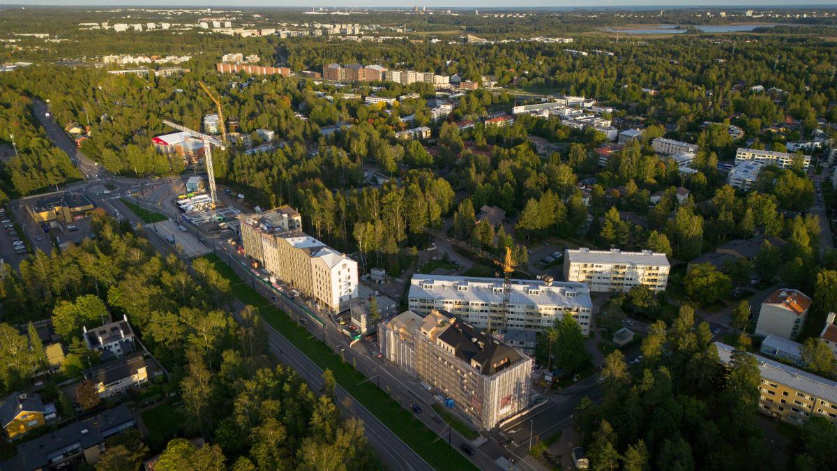 Flygbild över en grön stadsdel med flerbostadshus, en byggarbetsplats och omgivande skog.