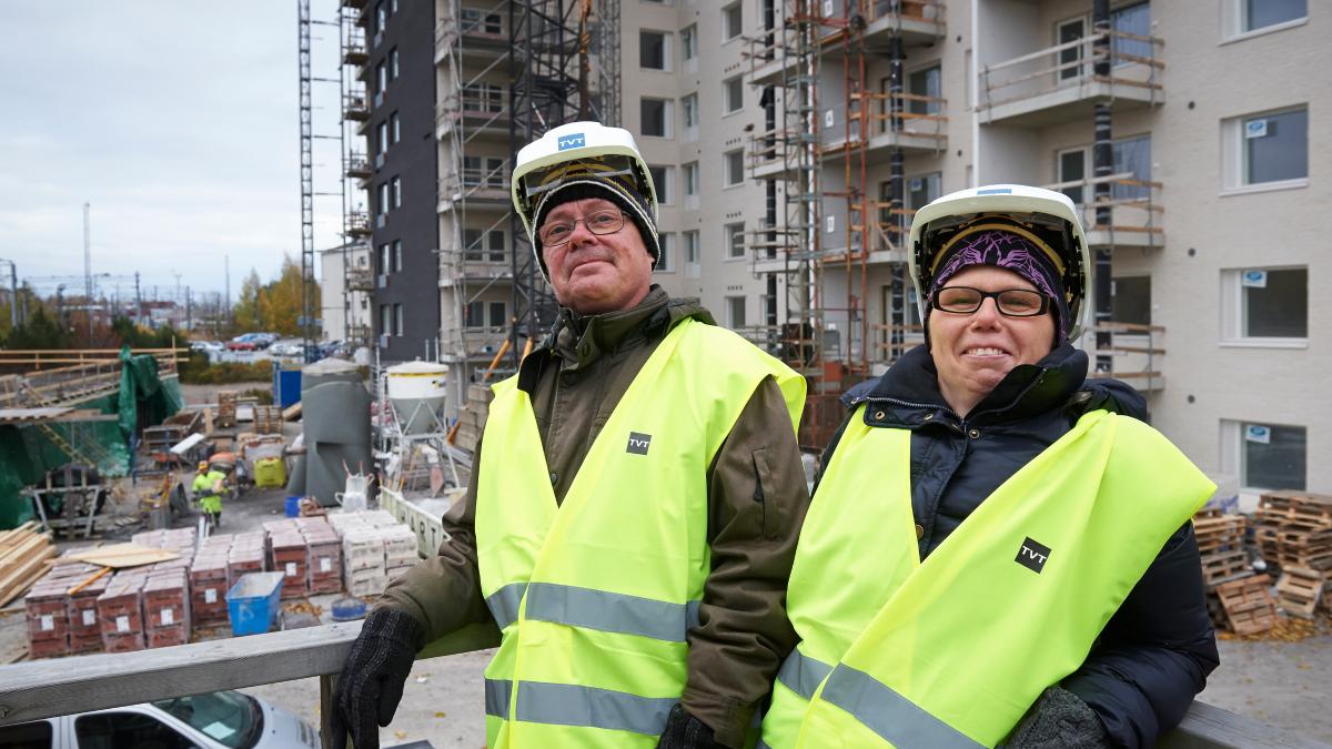 Kaksi henkilöä seisoo rakennustyömaalla, yllään keltaiset turvaliivit ja suojakypärät, taustalla kerrostalojen rakentaminen ja rakennusmateriaaleja.