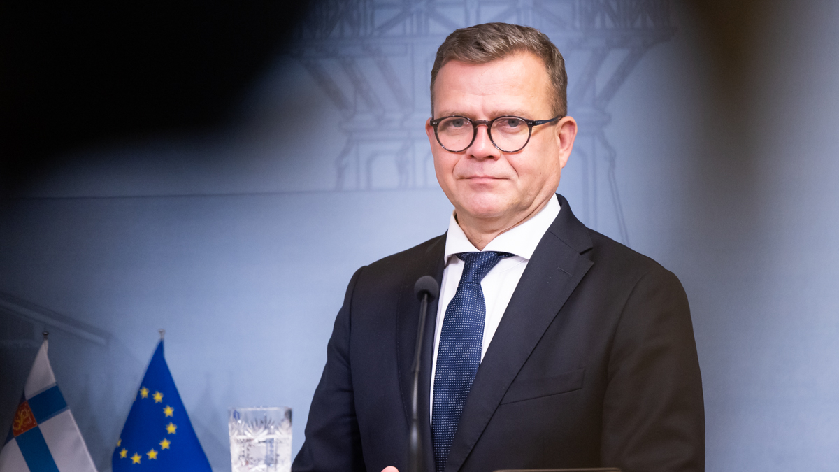 Pääministeri Orpo tiedotustilaisuudessa podiumin takana ja katsoo suoraan kameraan