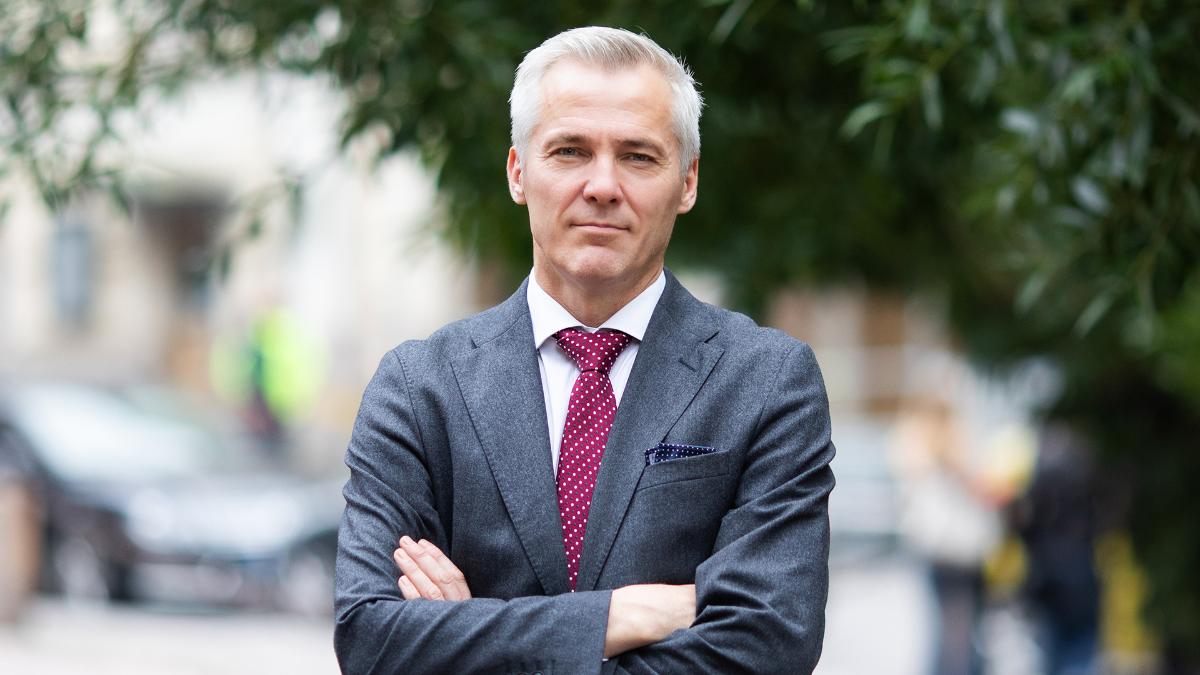 Ministeri Adlercreutz seisoo kädet puuskassa ja katsoo kameraan. Taustalla vihertävä puu.