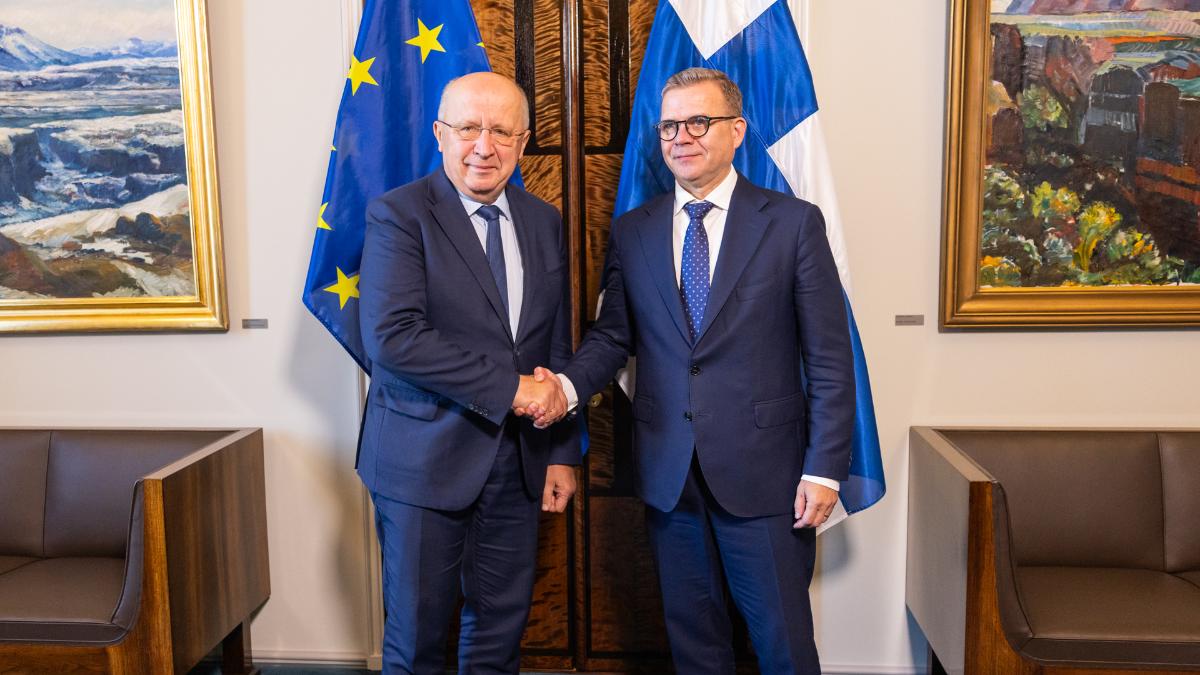 Pääministeri ja komissaari kättelevät EU-lipun ja Suomen lipun edessä.