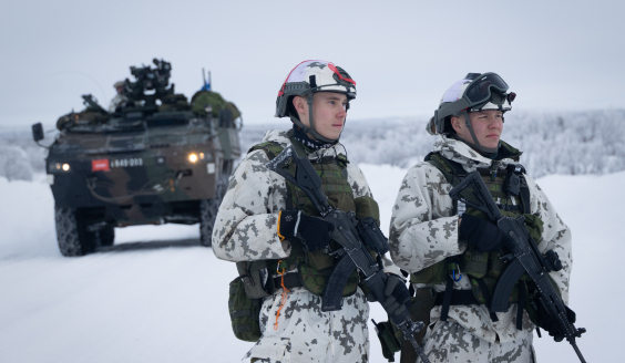 Two conscripts standing in combat load in snowy terrain. In the background, armoured personnel carrier Patria XA-360.