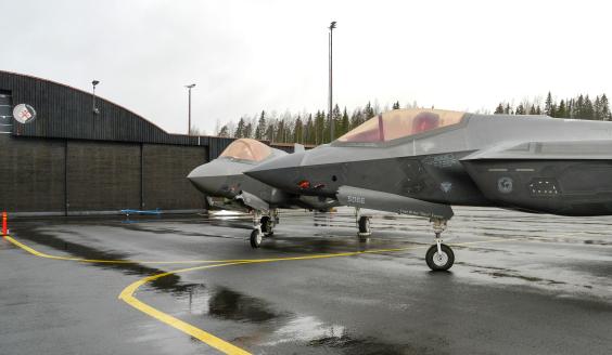 Kahden vierekkäin pysäköidyn yhdysvaltalaisen F-35A-hävittäjän keulat näkyvät kuvassa, joka on otettu Satakunnan lennostossa platta-alueella. Taivas on pilvinen ja asfaltti kostea. Taustalla, kuvan vasemmassa reunassa näkyvän hallin seinällä on Ilmataistelukeskuksen tunnus. 