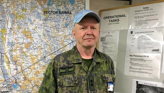 Maastopukuinen Sihvola seisoo karttaseinän edessä sininen YK:n lippalakki päässään