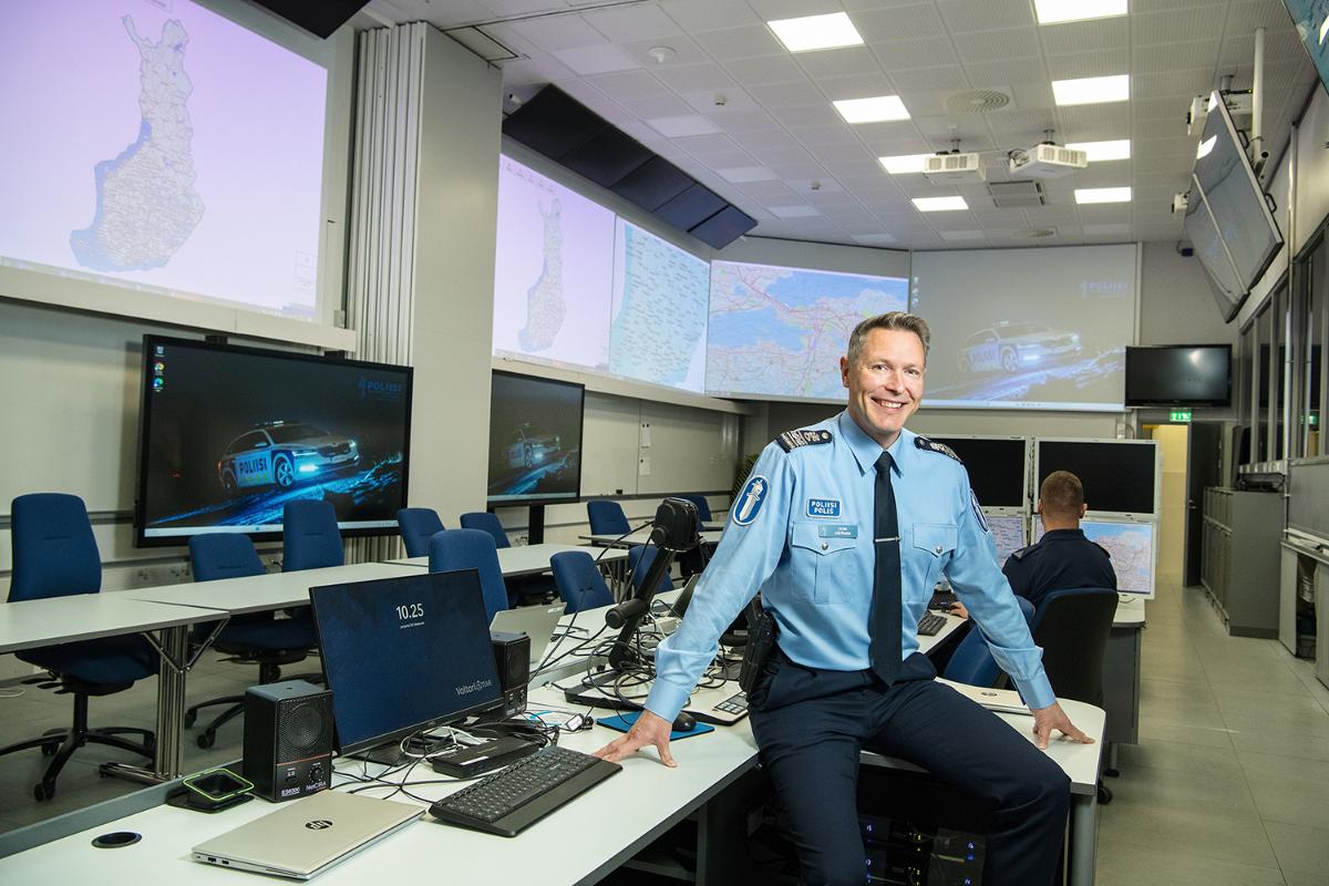 In the picture, a policeman is sitting on the edge of a table. In the background, computers and television screens are visible.