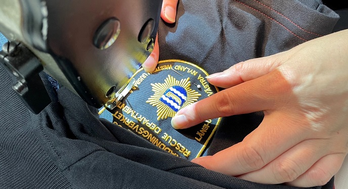 Lähikuva, jossa ompelukoneella ommellaan pelastuslaitoksen hihamerkkiä kiinni mustaan työpaitaan.