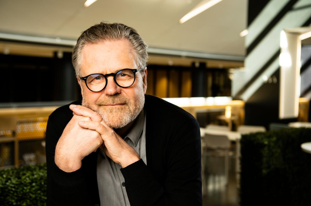 Kuvassa on mies, jolla on harmaat hiukset ja parta. Hänellä on pyöreät silmälasit ja päällään tummanvärinen neuletakki sekä harmaa kauluspaita. Hän nojaa käsiinsä, jotka on asetettu leuan alle, ja katsoo suoraan kameraan. Taustalla näkyy modernin toimiston sisätila, jossa on valaistuja elementtejä ja huonekaluja, kuten pöytiä ja tuoleja. Valo tulee osittain ylhäältä, luoden pehmeitä varjoja ympäristöön.